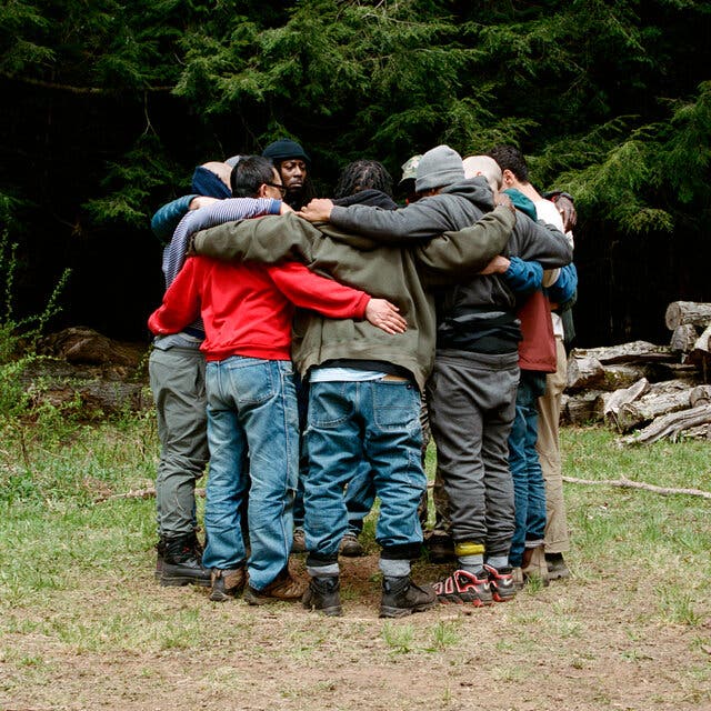 A group of men, mostly in jeans and sweatshirts, hug each other in a circle in the middle of a wooded area.